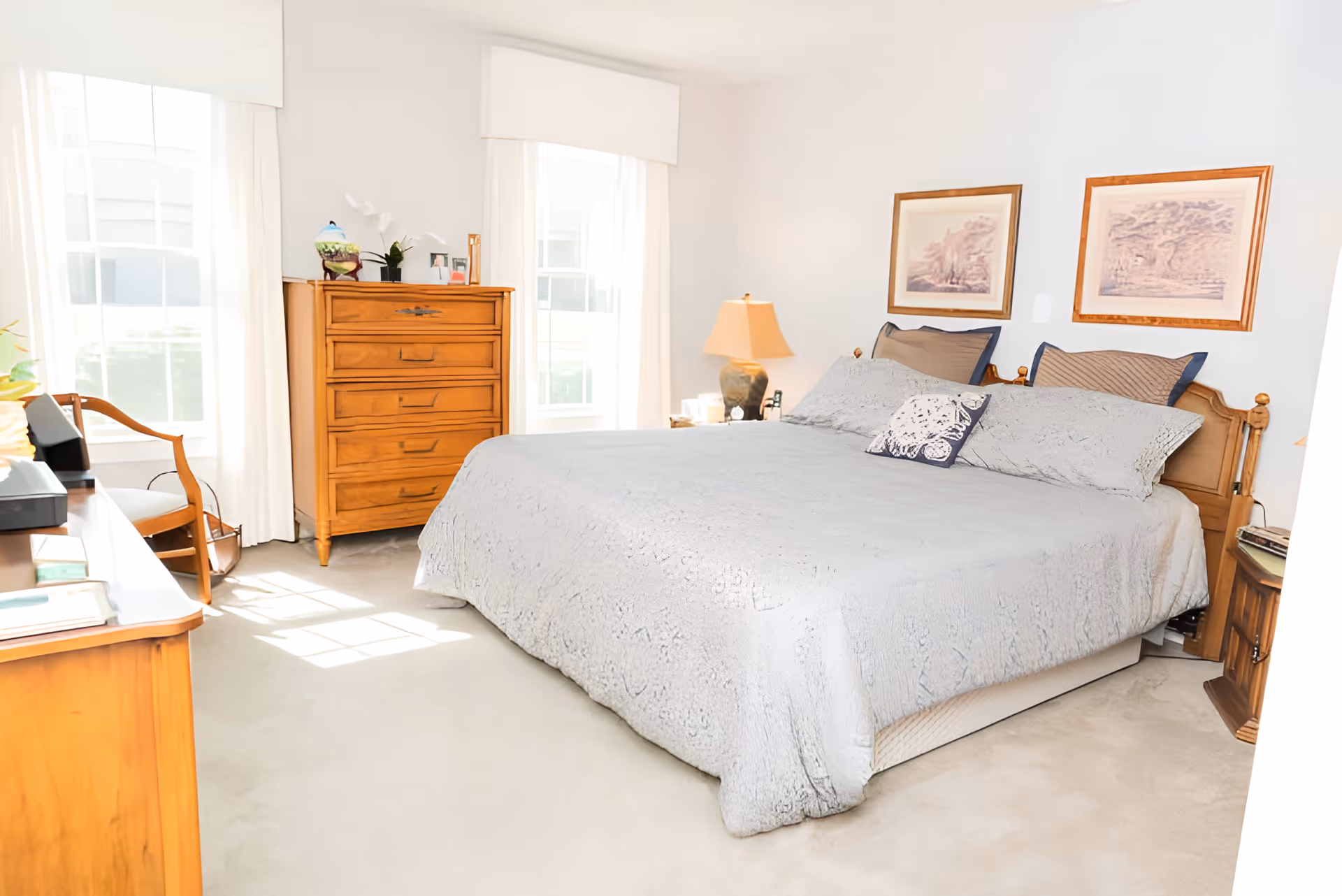 A bright bedroom with a large bed covered in a light gray quilt and multiple pillows. There are two framed pictures hanging above the bed. To the left of the bed is a wooden dresser with decorative items on top, and next to it are two tall windows with white curtains. A wooden chair is positioned near the dresser, and a bedside table with a lamp is on the right side of the bed. The room has light-colored walls and carpeted flooring.