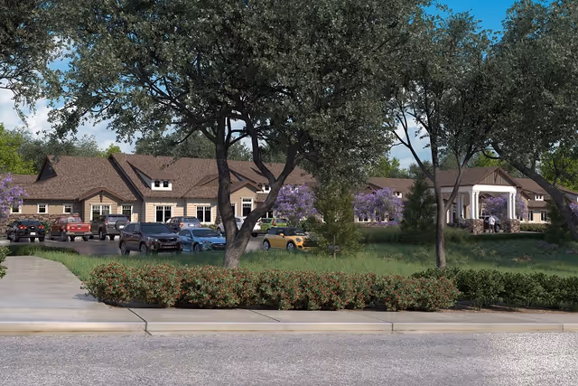 Exterior view of The Pavilion at El Dorado Hills senior living facility showing a single-story building with a brown roof surrounded by trees and bushes. Several cars are parked in front of the building, and a covered entrance is visible on the right side.