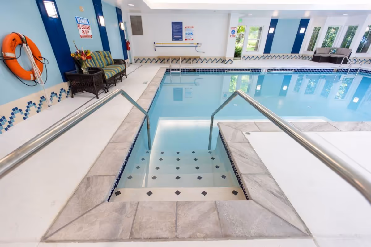 Indoor swimming pool with steps leading into the water, stainless steel handrails, and tiled edges. The pool area has blue and white walls with safety signs, a life preserver mounted on the wall, and seating including a striped cushioned bench and a wicker table with flowers. Large windows allow natural light into the space.
