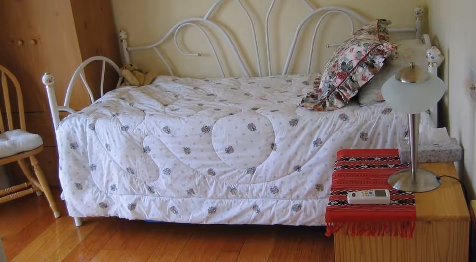 A white metal-framed single bed with floral bedding and pillows beside a wooden nightstand holding a lamp and a remote.