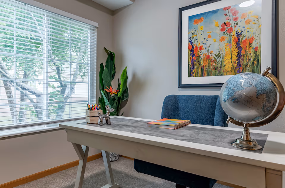 A bright office or study area with a white desk featuring a globe, a colorful notebook, and a small container holding pens and pencils. Behind the desk is a blue upholstered chair. A large window with white blinds lets in natural light, and a tall green plant with orange flowers is placed in the corner. A framed painting of colorful flowers hangs on the wall above the chair.