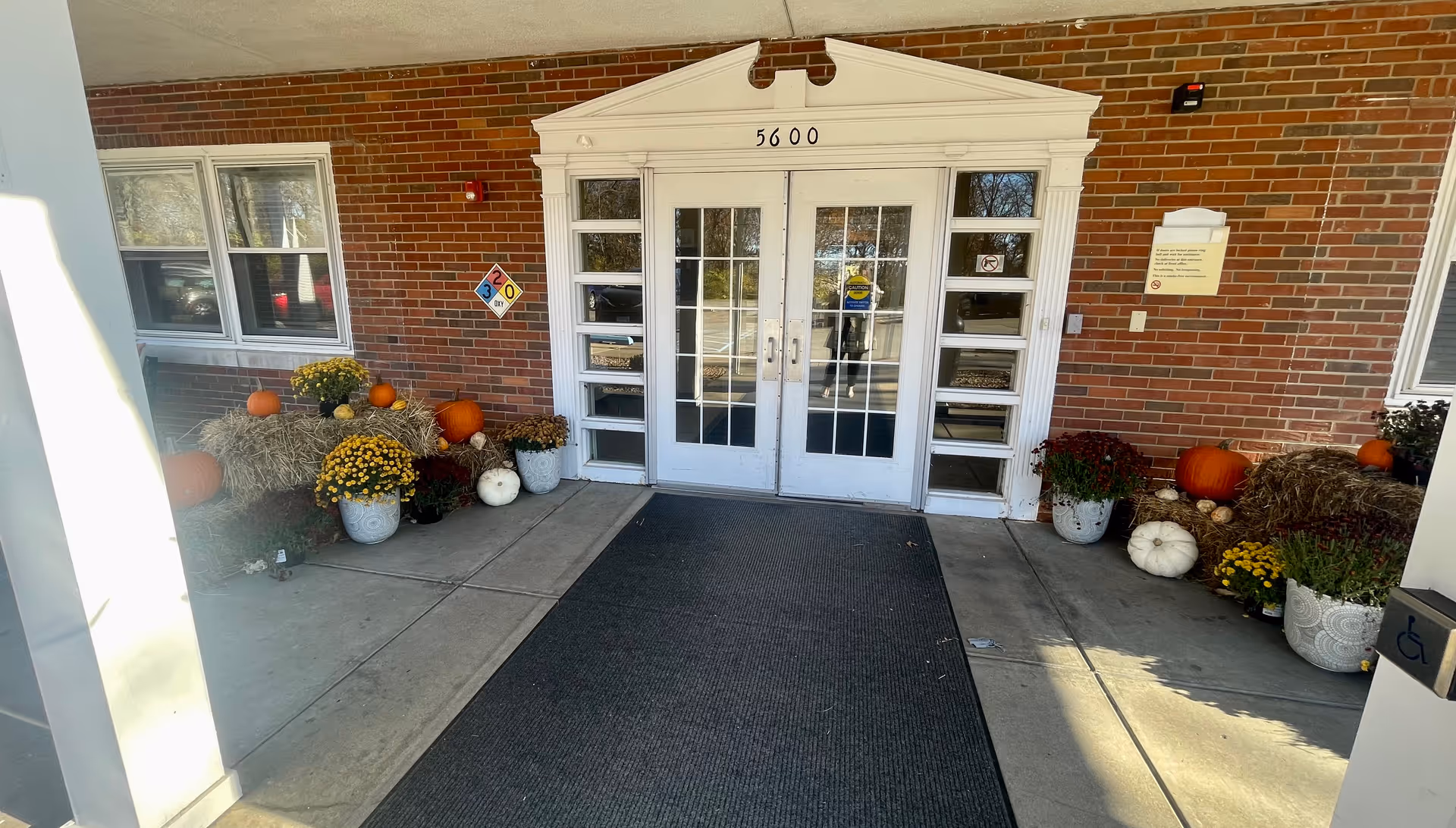 Entrance to a building with double glass doors framed by white trim and the number 5600 above. The entrance is decorated with pumpkins, hay bales, and potted flowers on both sides. The building exterior is brick, and there is a black mat on the concrete floor leading to the doors.