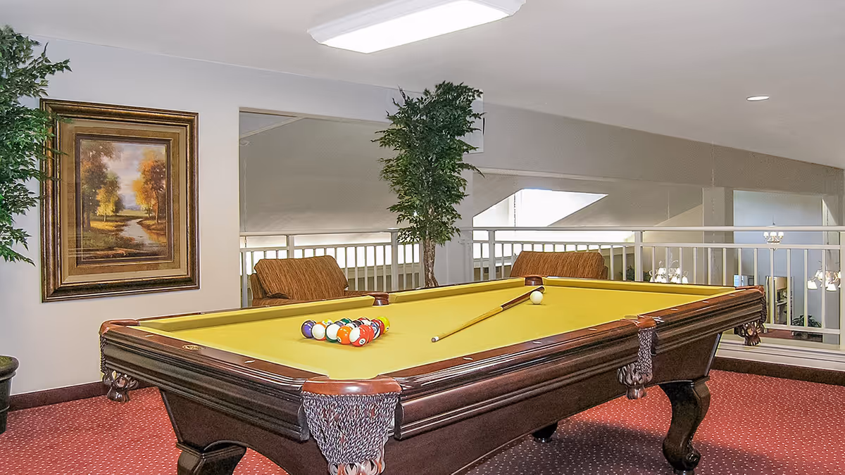 Indoor recreational area with a yellow felt pool table set up for a game, two brown armchairs, a large framed painting of a landscape on the wall, potted plants, and a railing overlooking a lower level with hanging lights.