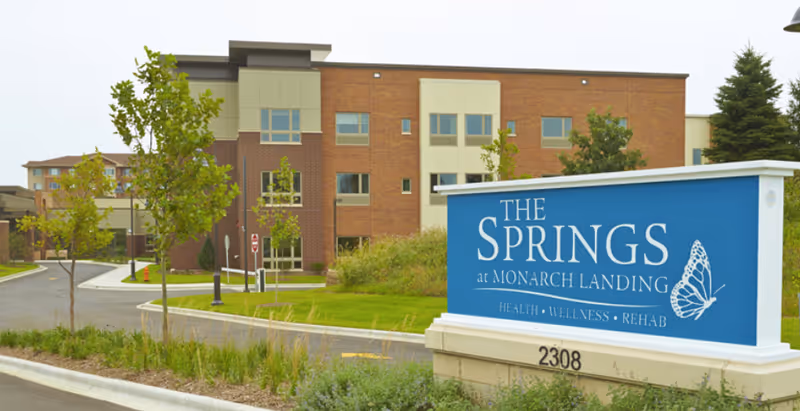 Exterior view of The Springs at Monarch Landing senior living facility showing a modern brick building with multiple windows, a curved driveway, landscaped greenery, and a large blue sign with the facility's name and services including health, wellness, and rehab.