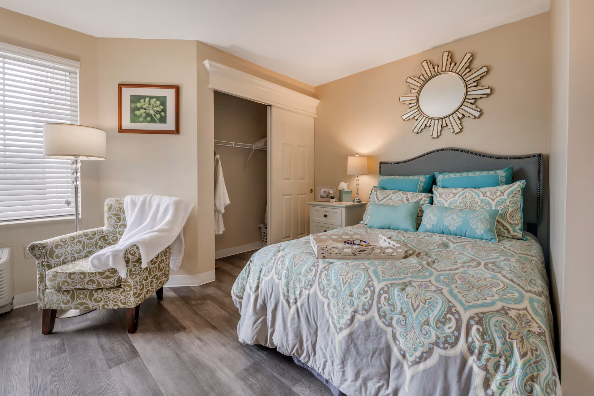 Bright furnished bedroom with a teal-and-gray patterned bed and multiple pillows, a sunburst mirror above the headboard, an upholstered armchair by a window, and an open closet.