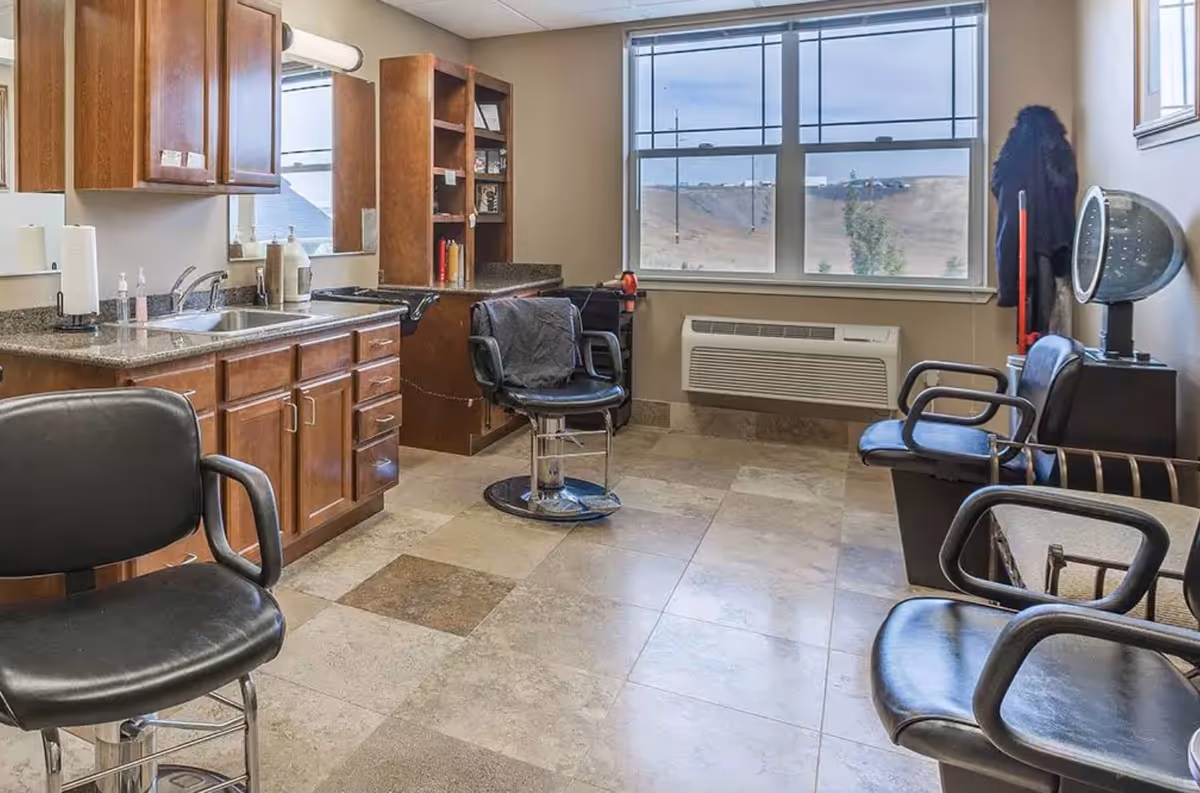 A small salon room with multiple styling chairs, a shampoo sink, cabinetry, and a hooded hair dryer under a large window.