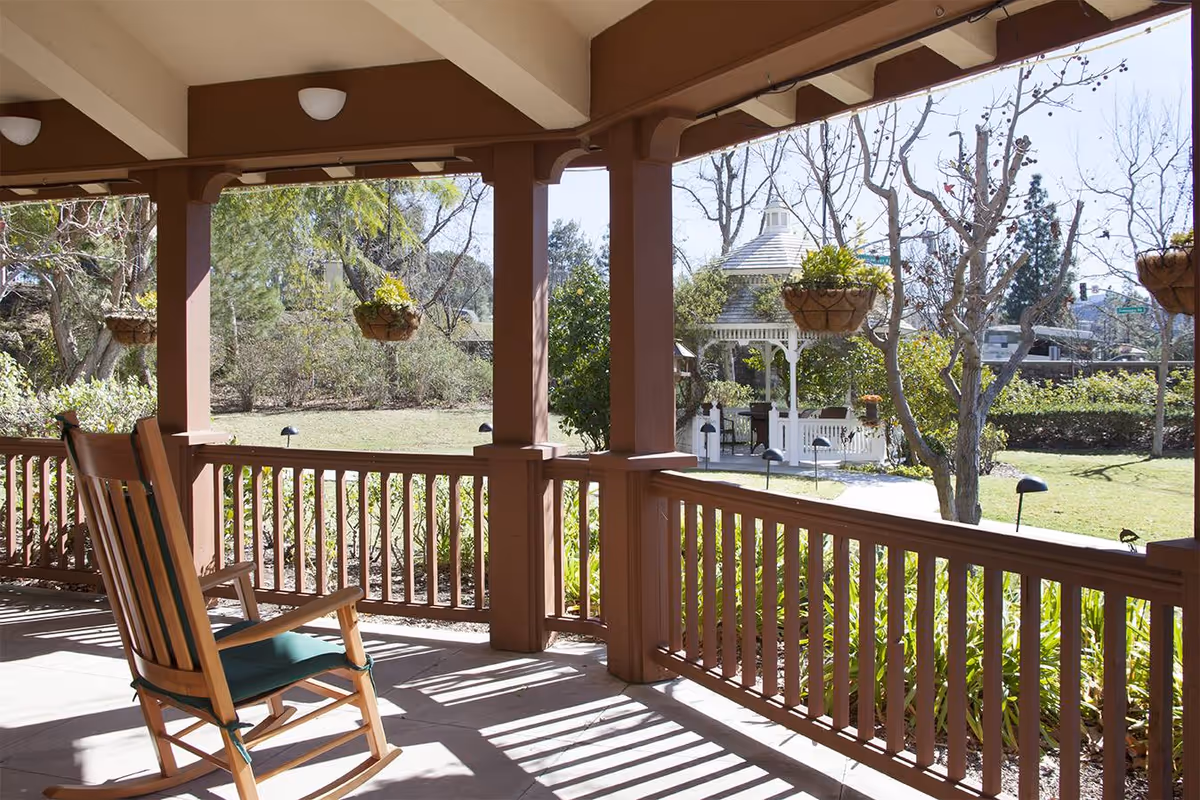 Covered wooden porch with a rocking chair, hanging planters, and a view of a gazebo and garden.