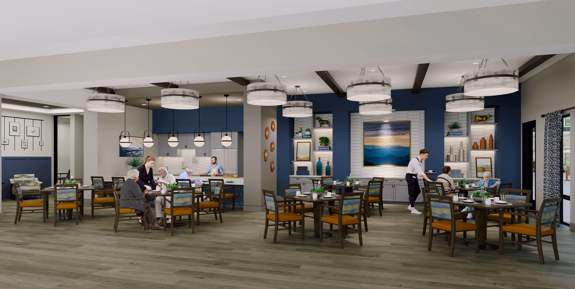 A spacious dining room in a senior living facility with several wooden tables and chairs. Residents are seated at tables being attended by staff. The room features modern pendant and ceiling lights, blue accent walls with decorative shelves, and a kitchen area in the background.