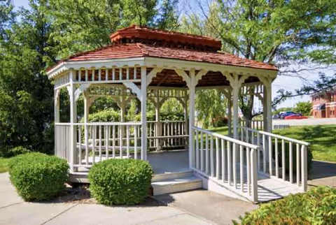 White wooden gazebo with red roof sits amid green bushes and trees in a sunny park.