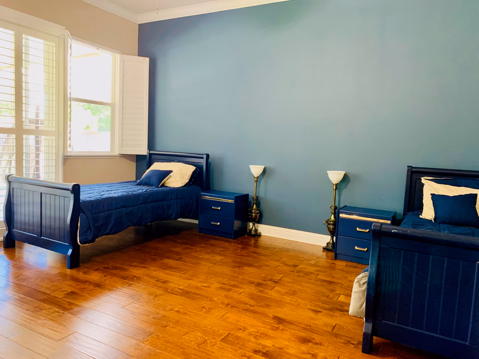 A bedroom with two single beds featuring dark blue frames and matching bedding. Each bed has a pillow and a cushion. Between the beds are two blue nightstands with brass handles, each accompanied by a brass floor lamp with a white lampshade. The room has wooden flooring and a blue accent wall. There is a window with white plantation shutters allowing natural light into the room.
