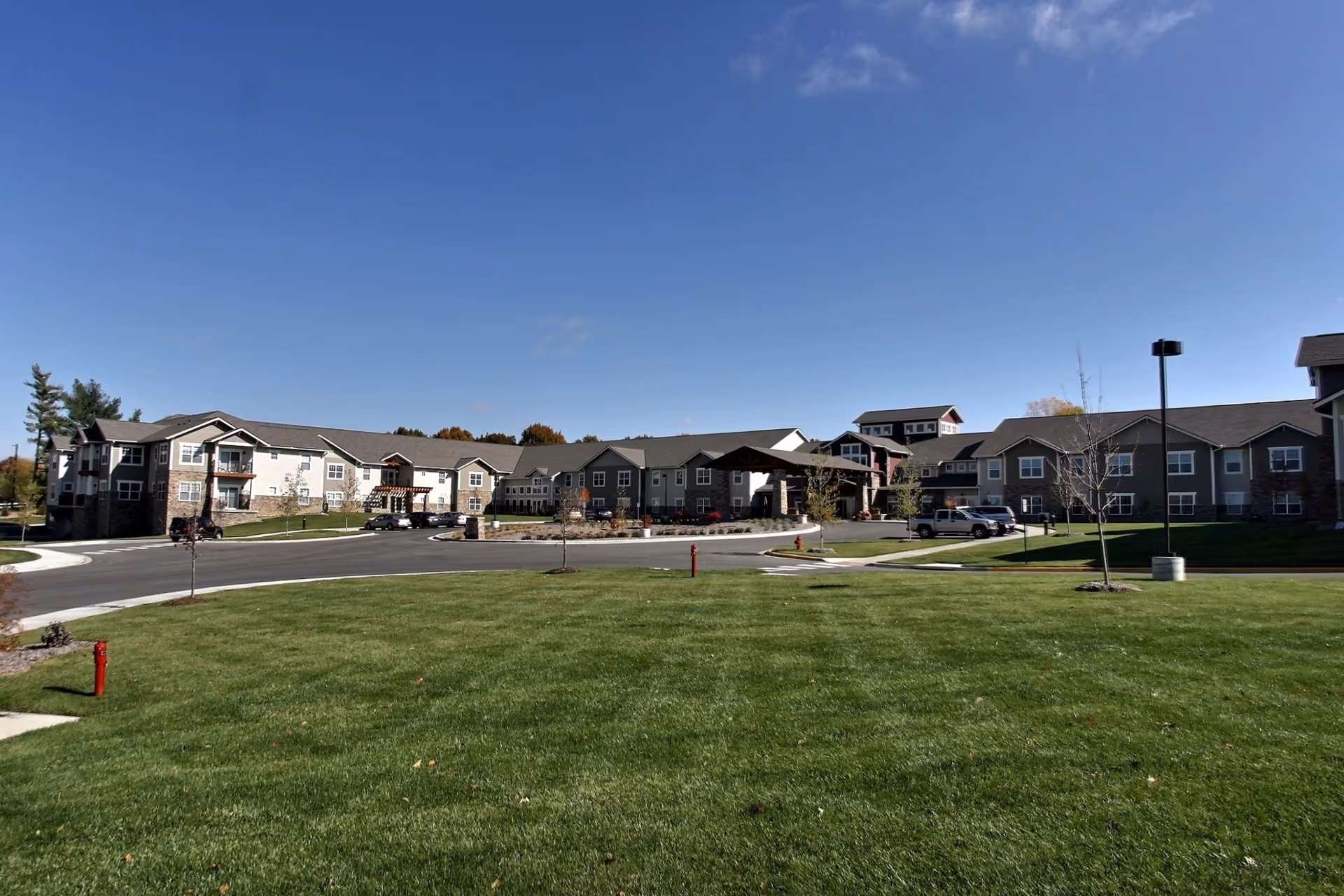 Front exterior of a senior living building with a circular driveway, parked cars and a large green lawn under a clear blue sky.