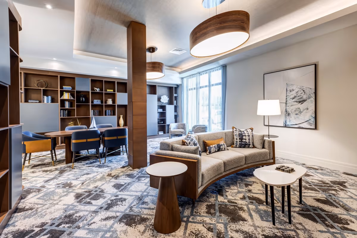 A modern and stylish living room area in a senior living facility featuring a curved beige sofa with patterned cushions, two small white and wooden side tables, a floor lamp, and abstract wall art. In the background, there is a wooden dining table with blue and mustard chairs, built-in wooden shelves with decorative items and books, large windows with sheer curtains, and two large circular ceiling lights.