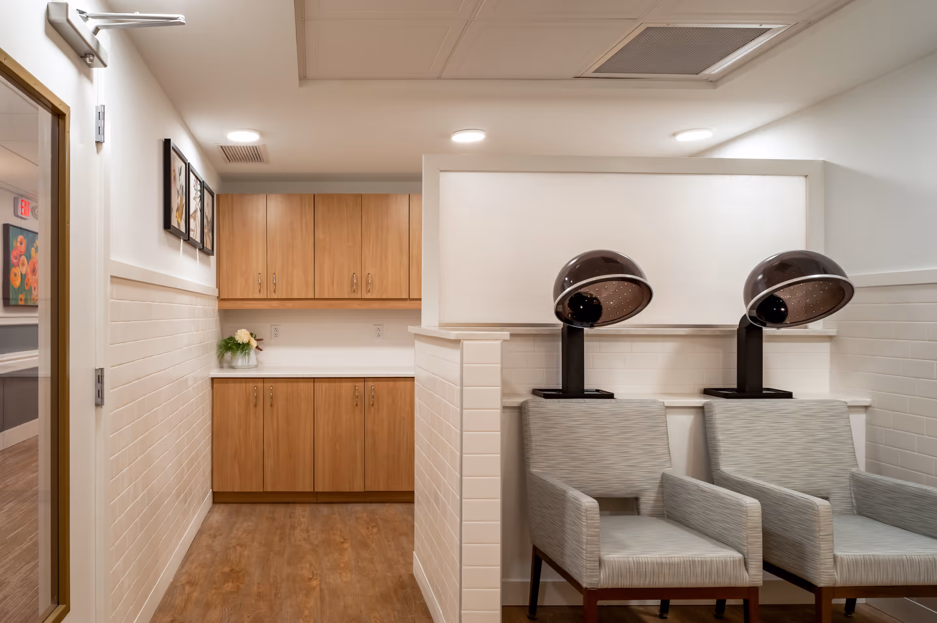 Small salon area with two hooded hair dryers over upholstered chairs, wood cabinets, and a tiled half-wall.