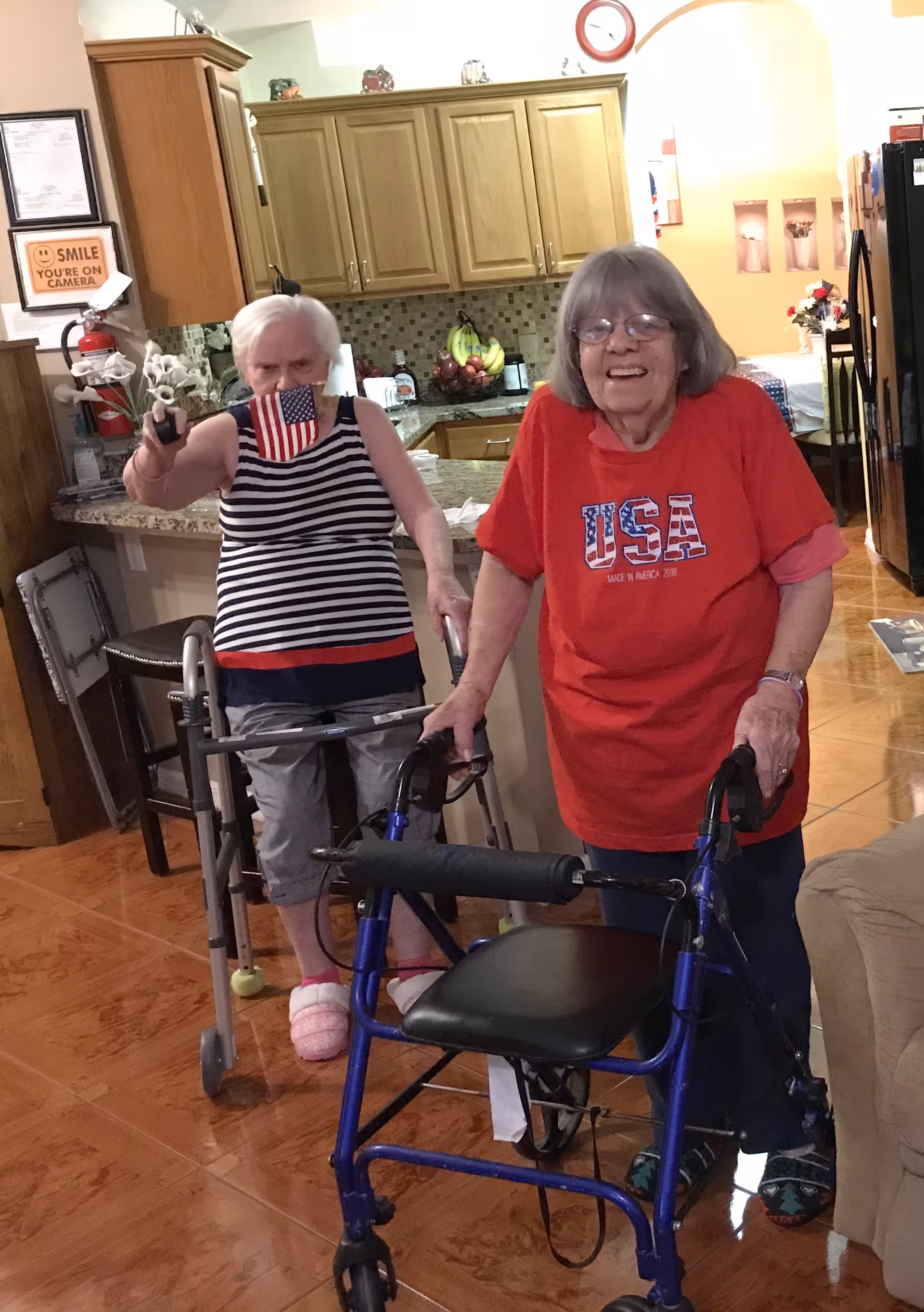 Two elderly women standing in a kitchen area, each using a walker. One woman is wearing a red USA t-shirt and glasses, smiling at the camera. The other woman is wearing a striped tank top and holding a small American flag and a small bouquet of flowers. The kitchen has wooden cabinets, a fruit bowl, and various items on the counter.