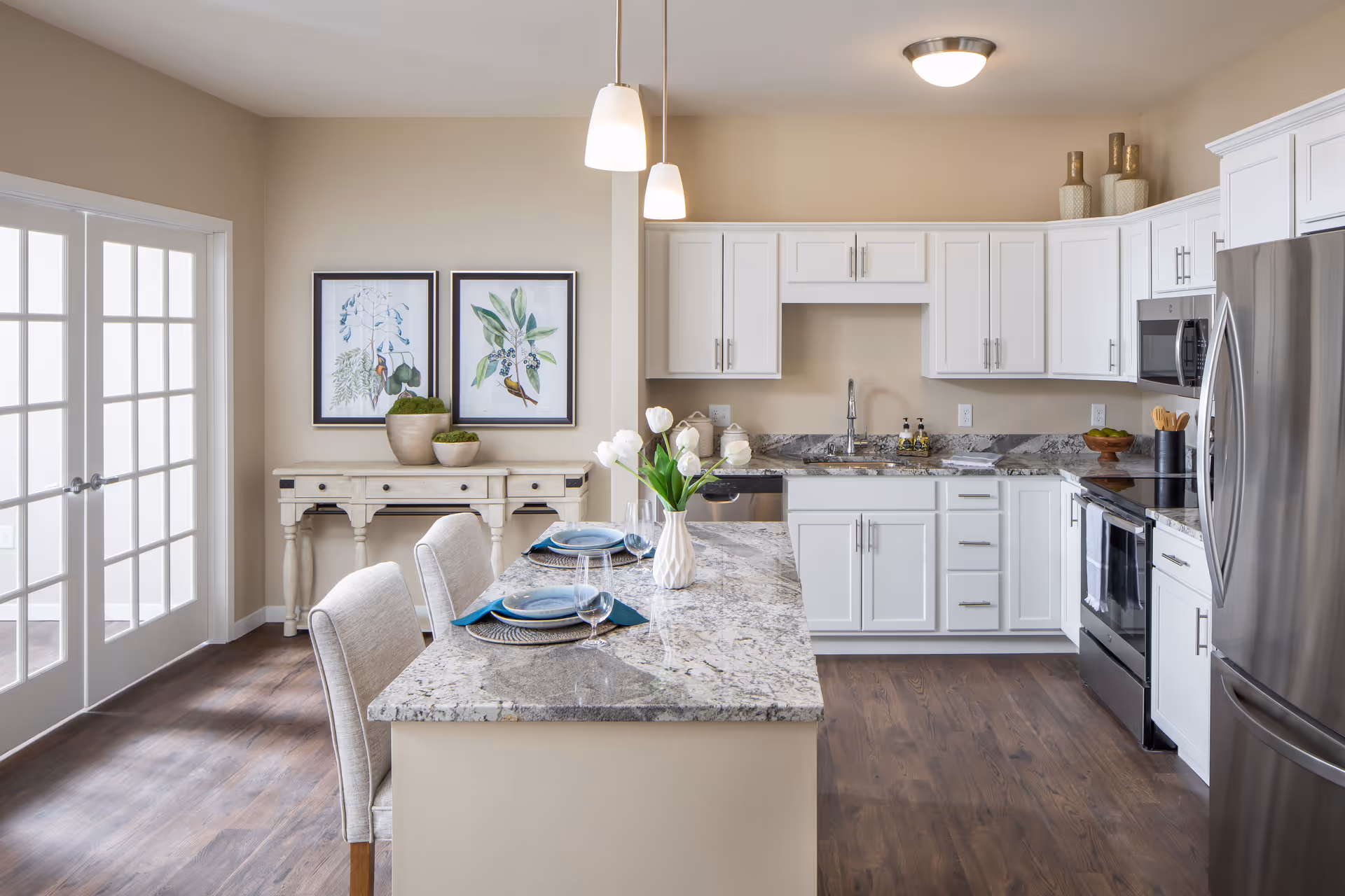 Bright modern kitchen with white cabinets, granite countertops, stainless steel appliances, and a kitchen island set with plates, glasses, and napkins. Two pendant lights hang above the island. There is a wooden console table against the wall with two framed botanical prints above it and decorative pots on top. The floor is wood, and there are French doors to the left.