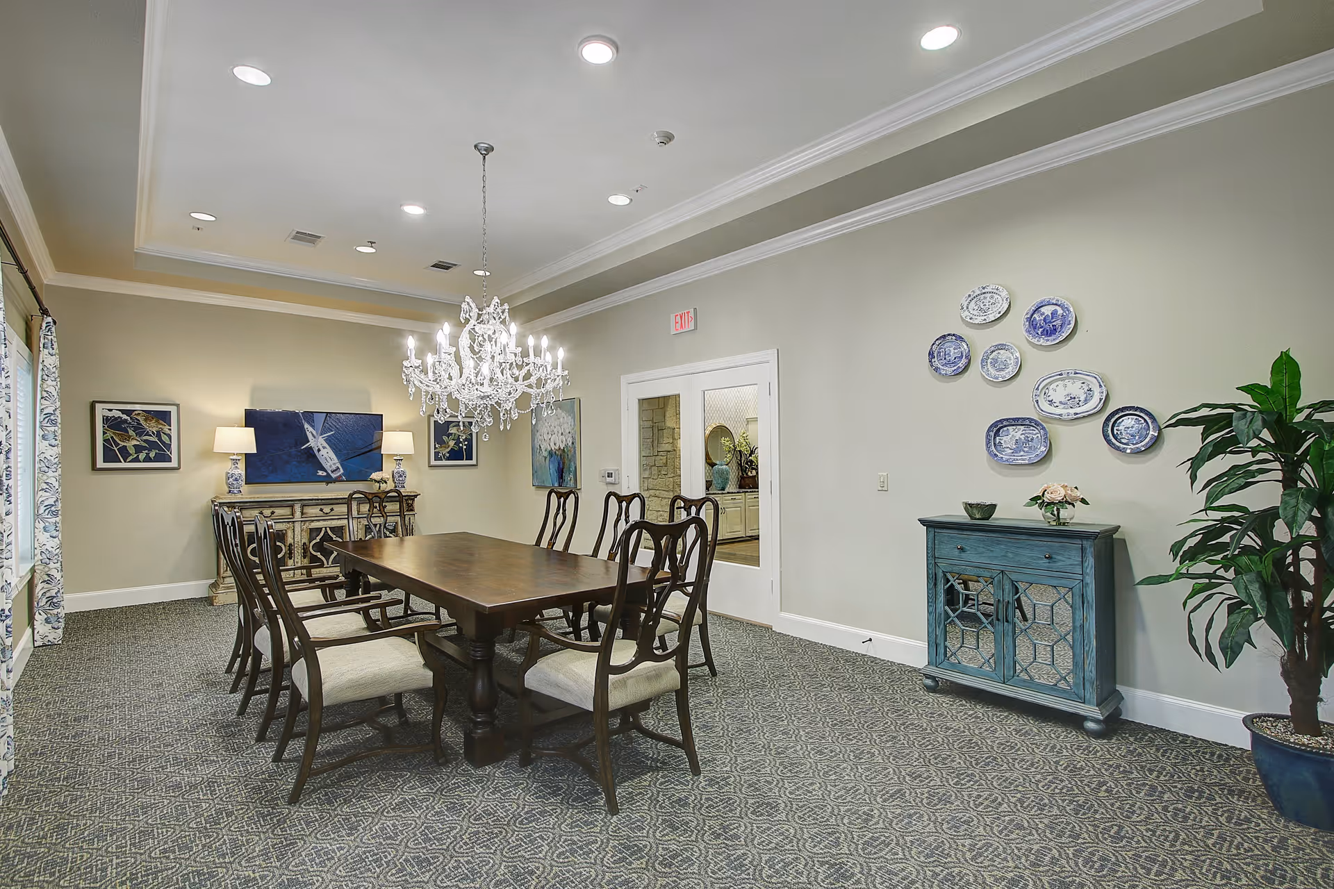 A formal dining room with a long wooden table surrounded by eight cushioned chairs. A crystal chandelier hangs above the table. The room has patterned carpet flooring, beige walls, and white trim. On one wall, there is a blue cabinet with decorative plates arranged above it and a potted plant beside it. On the opposite wall, there is a sideboard with two lamps and framed artwork above it. A glass door leads to another room.