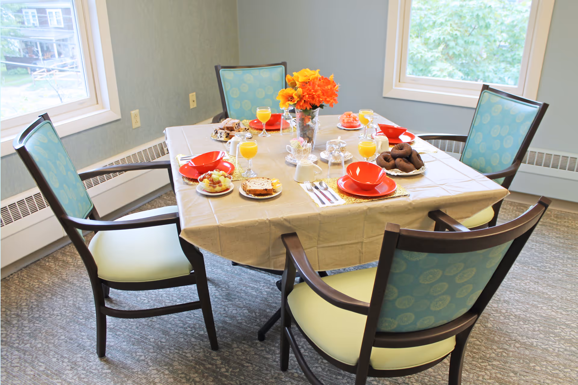 A dining table set for four with a beige tablecloth, red bowls and plates, glasses of orange juice, plates of pastries, fruit, and donuts, and a vase with orange and yellow flowers in a room with two windows and four chairs with blue patterned backs and light green seats.