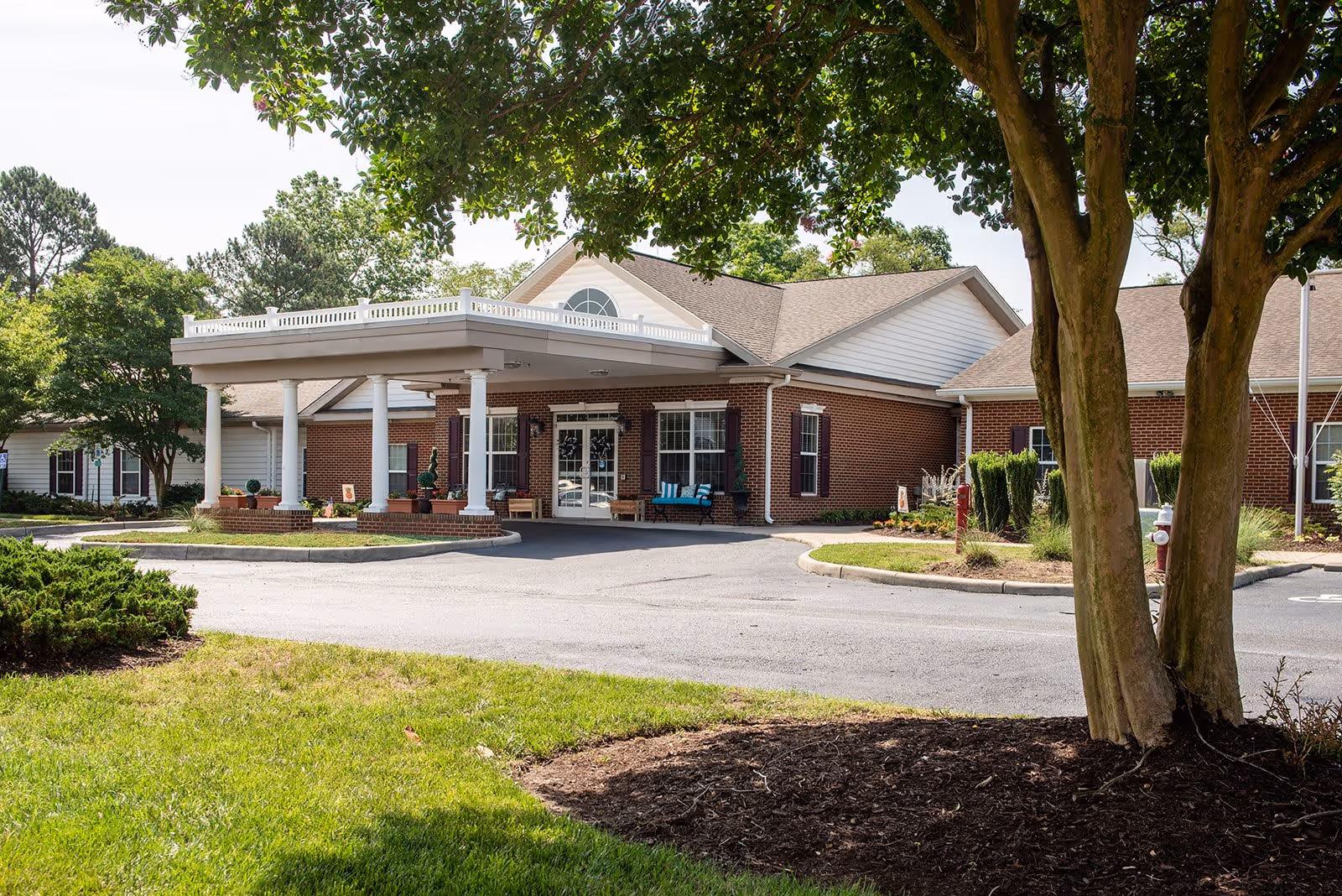 Exterior view of a single-story brick building with white trim and a covered entrance supported by white columns. There are benches near the entrance and trees and greenery surrounding the driveway and lawn area.