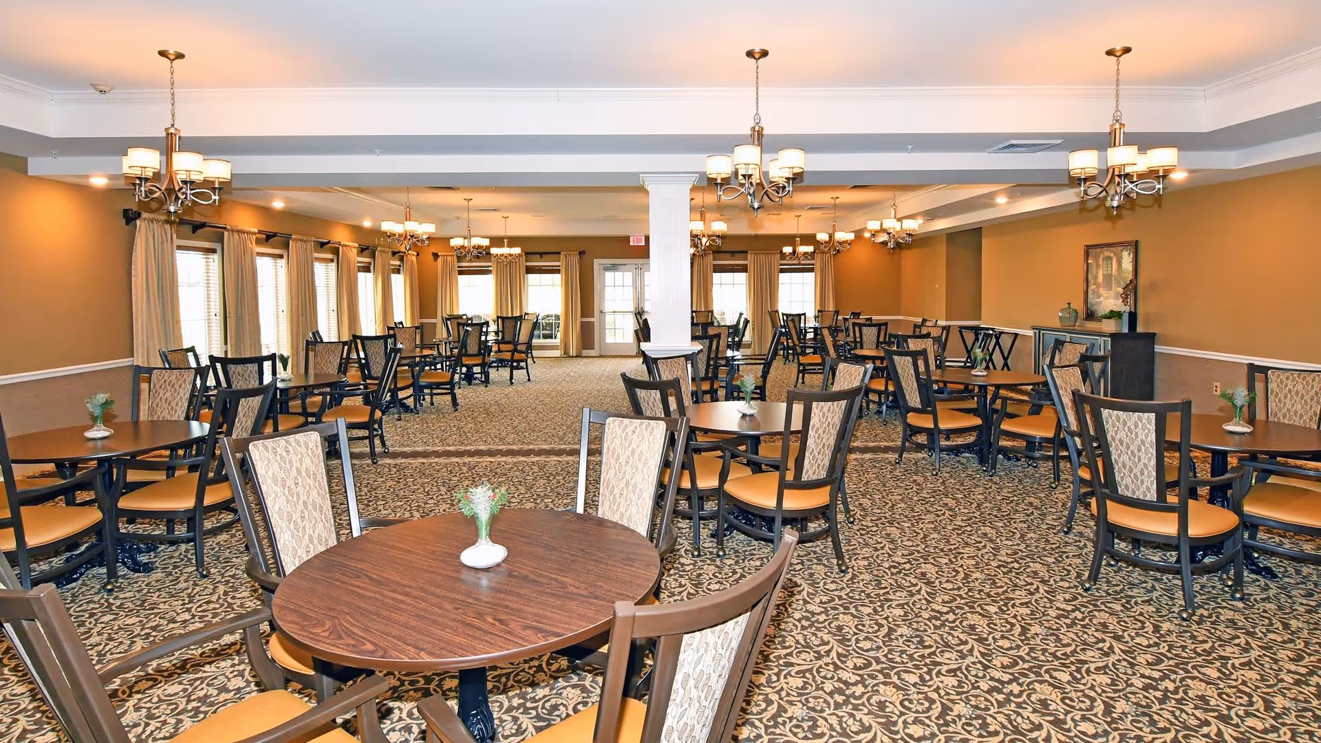 A spacious dining room with multiple round wooden tables and cushioned chairs arranged neatly. The room features patterned carpet flooring, beige walls, large windows with curtains, and several chandeliers hanging from the ceiling providing warm lighting.
