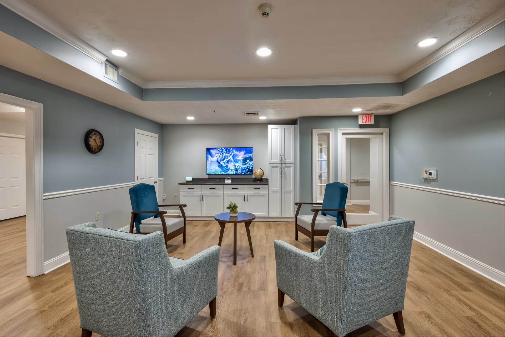 A cozy common area with four armchairs arranged around a small round wooden table. The room has light blue walls with white trim, wood flooring, and recessed ceiling lights. A flat-screen TV displaying an underwater scene is mounted on a white cabinet against the far wall. There is a clock on the left wall and an exit sign above a door on the right side.