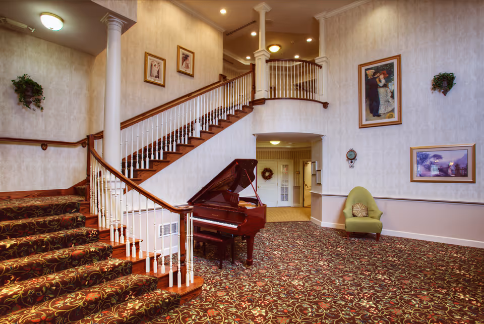 Interior view of a senior living facility lobby area with a grand piano, a carpeted staircase with white railings, a green armchair with a decorative pillow, and framed artwork on the walls.