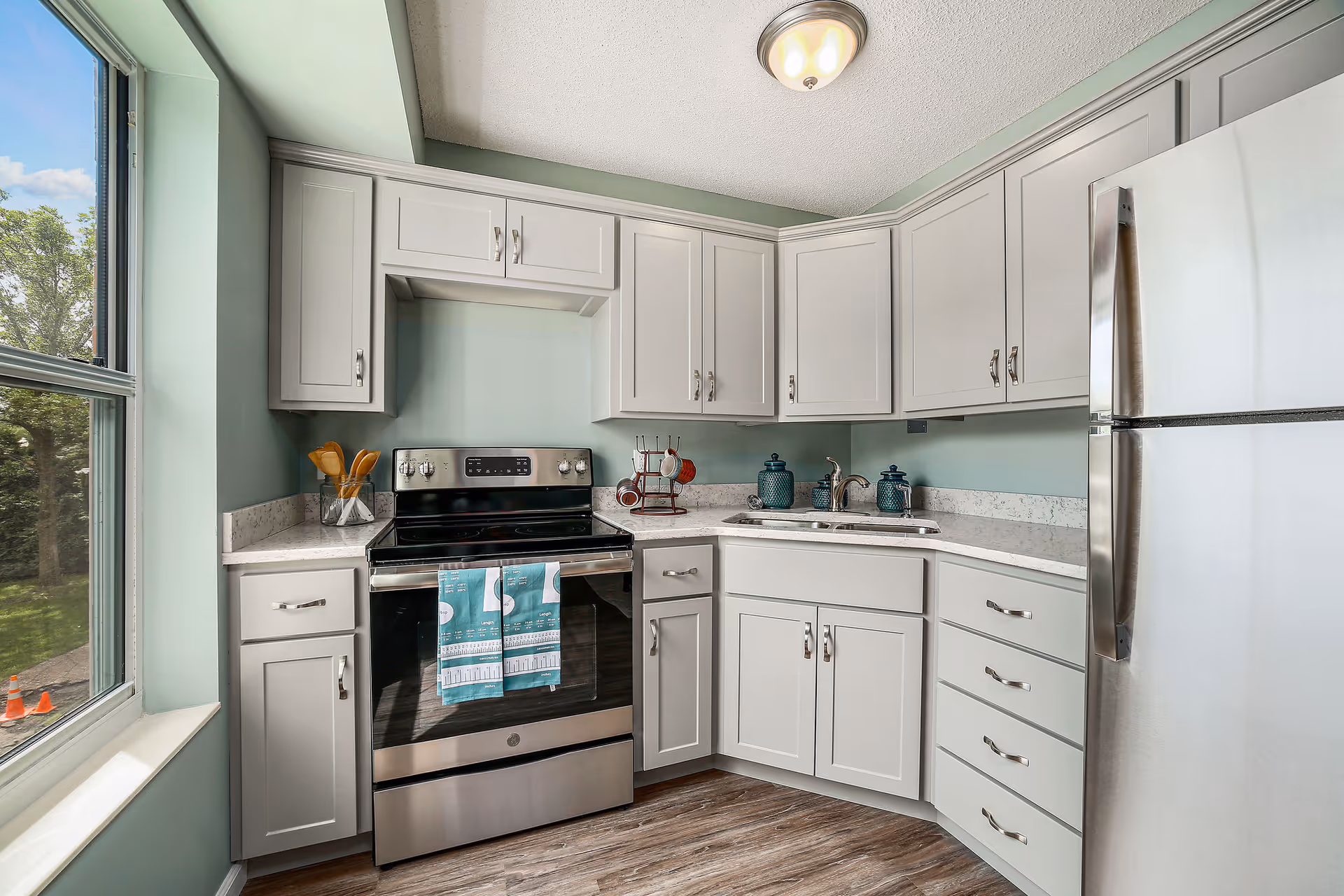 A modern kitchen with light gray cabinets, a stainless steel electric stove with a teal towel hanging on the handle, a stainless steel refrigerator, a double sink with two teal jars on the counter, and a large window showing trees outside. The walls are painted light green and the floor has wood-like vinyl planks.