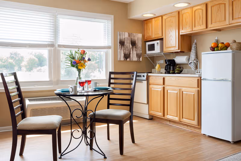 Sunlit kitchen and dining nook with a small round table and two chairs, wooden cabinets, stove and a compact refrigerator.