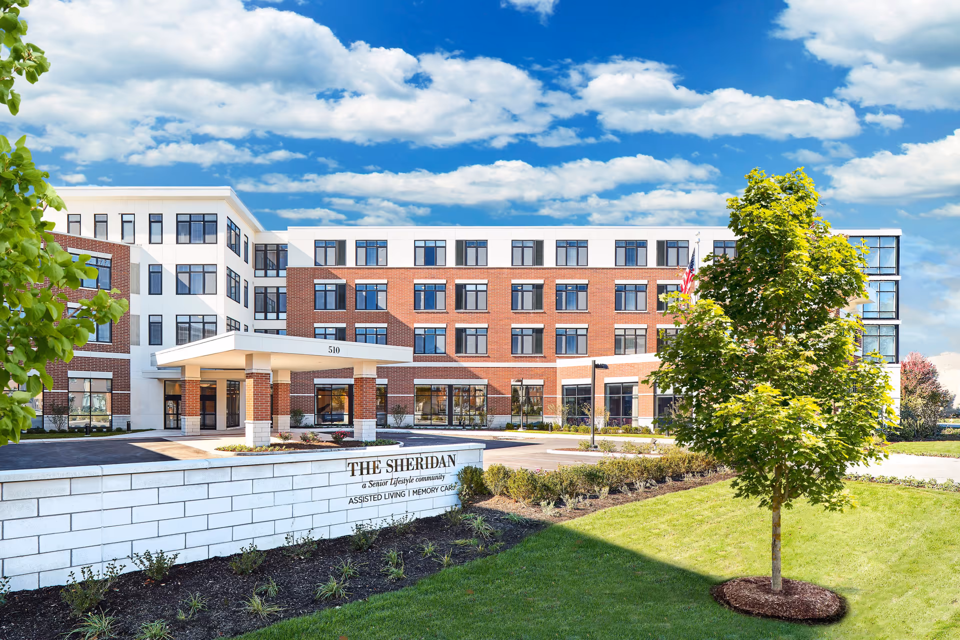 Exterior view of The Sheridan at Park Ridge senior living facility, showing a modern multi-story building with a brick and white facade, large windows, a covered entrance, landscaped greenery, and a tree in the foreground under a partly cloudy sky.