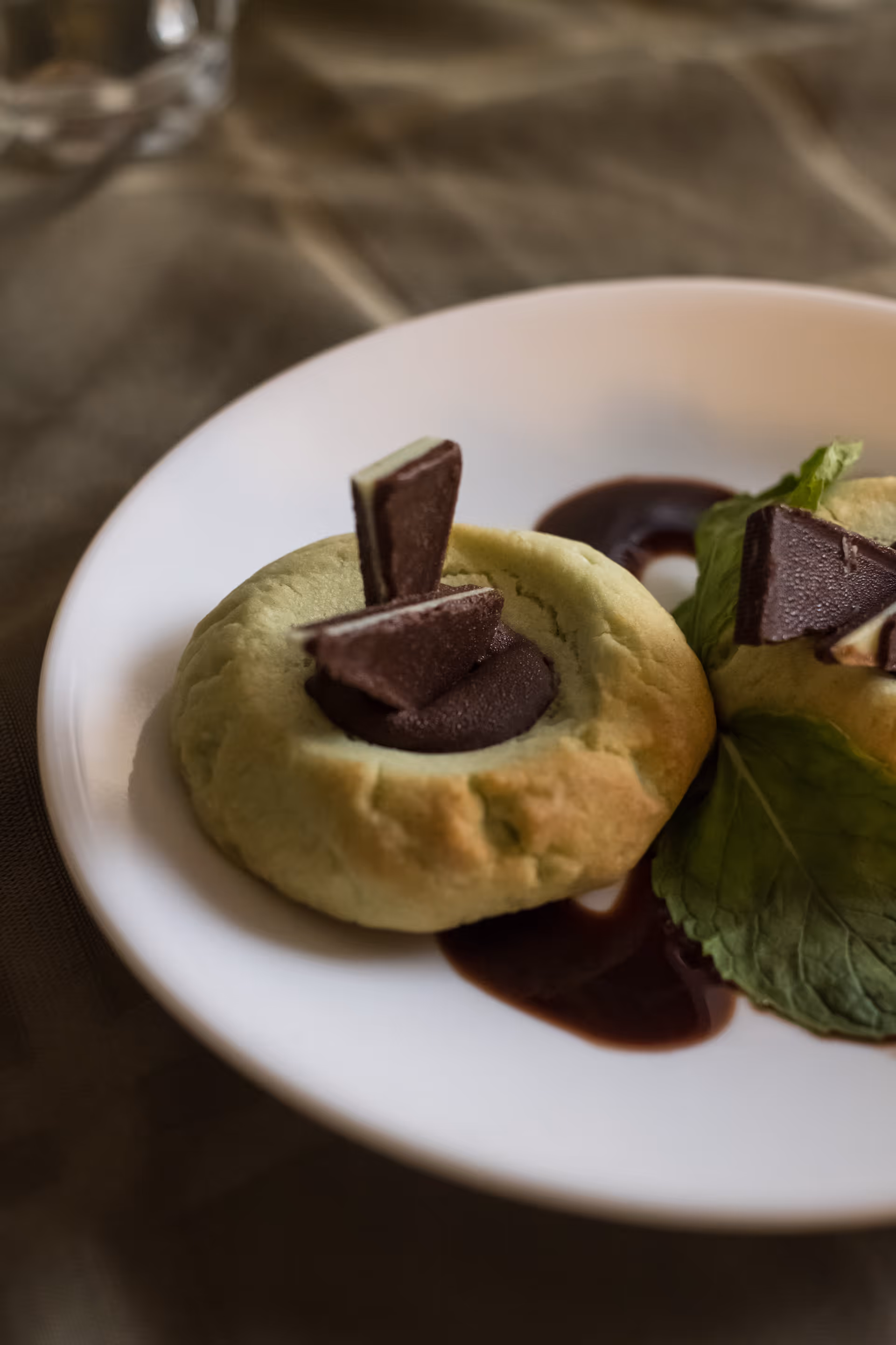 Close-up of two cookies on a white plate, each topped with a swirl of chocolate and two pieces of chocolate, garnished with a green leaf and a drizzle of chocolate sauce on the plate.