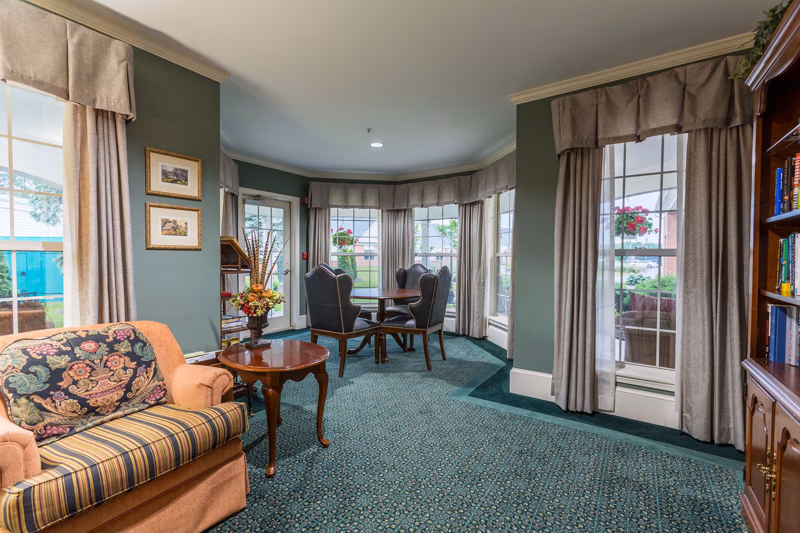 A cozy common area in a senior living facility featuring a cushioned armchair with a patterned pillow, a small wooden side table with a floral arrangement, a round wooden table with four upholstered chairs, large windows with beige curtains allowing natural light, and a wooden bookshelf filled with books.
