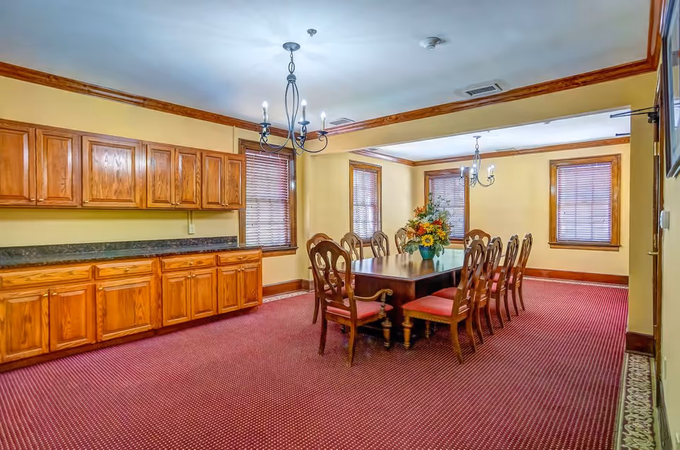 A dining room with a long wooden table surrounded by ten wooden chairs with red cushions. The room has yellow walls, three windows with wooden blinds, and wooden cabinets along one wall. Two chandeliers hang from the ceiling, and a floral arrangement is placed in the center of the table. The floor is covered with a red patterned carpet.