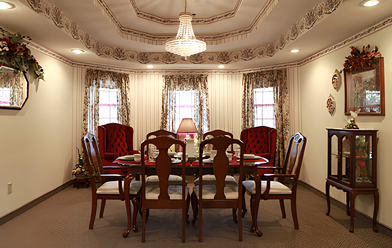 A formal dining room with a wooden dining table set for six, surrounded by six wooden chairs with cushioned seats. The room features three tall windows with floral curtains, a chandelier hanging from a decorative ceiling, and a small cabinet with a mirror and decorative items on the right wall.