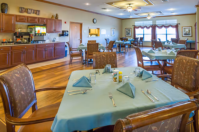 Dining room with several set tables covered in teal tablecloths, wooden chairs, and a serving counter along the wall.