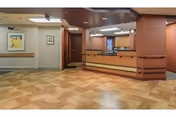 Interior view of a reception area in a senior living facility with a wooden reception desk, beige and brown tiled flooring, and walls painted in neutral tones. The area is well-lit with ceiling lights, and there are framed pictures on the walls.