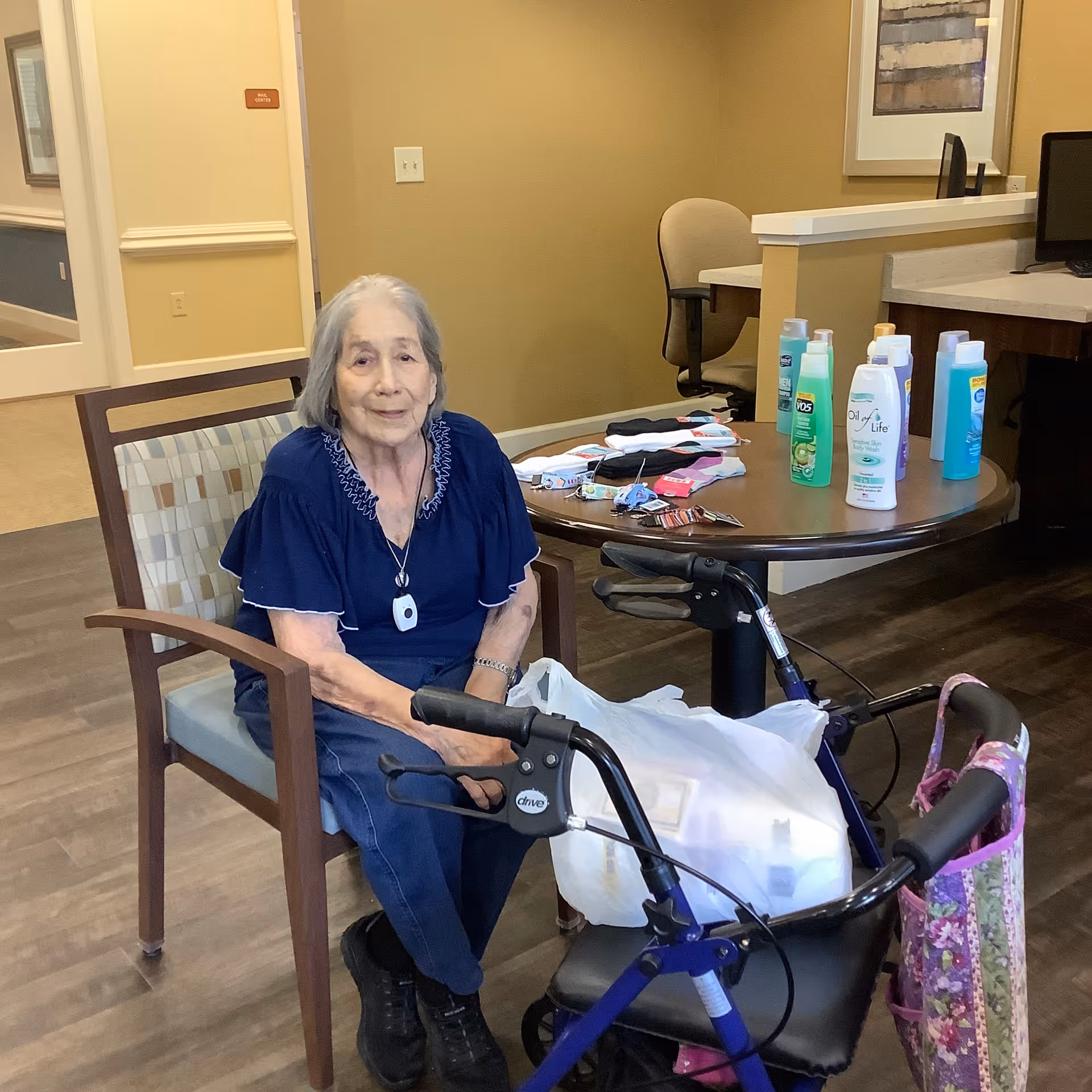 An elderly woman with gray hair wearing a navy blue blouse and dark pants is sitting on a wooden chair with a patterned cushion in a room. In front of her is a blue walker with a floral bag and a white plastic bag hanging from it. Behind her is a round table with various bottles of lotion and personal care items, as well as some folded clothes and small items. The room has beige walls, a wooden floor, and office furniture including a desk and chair in the background.