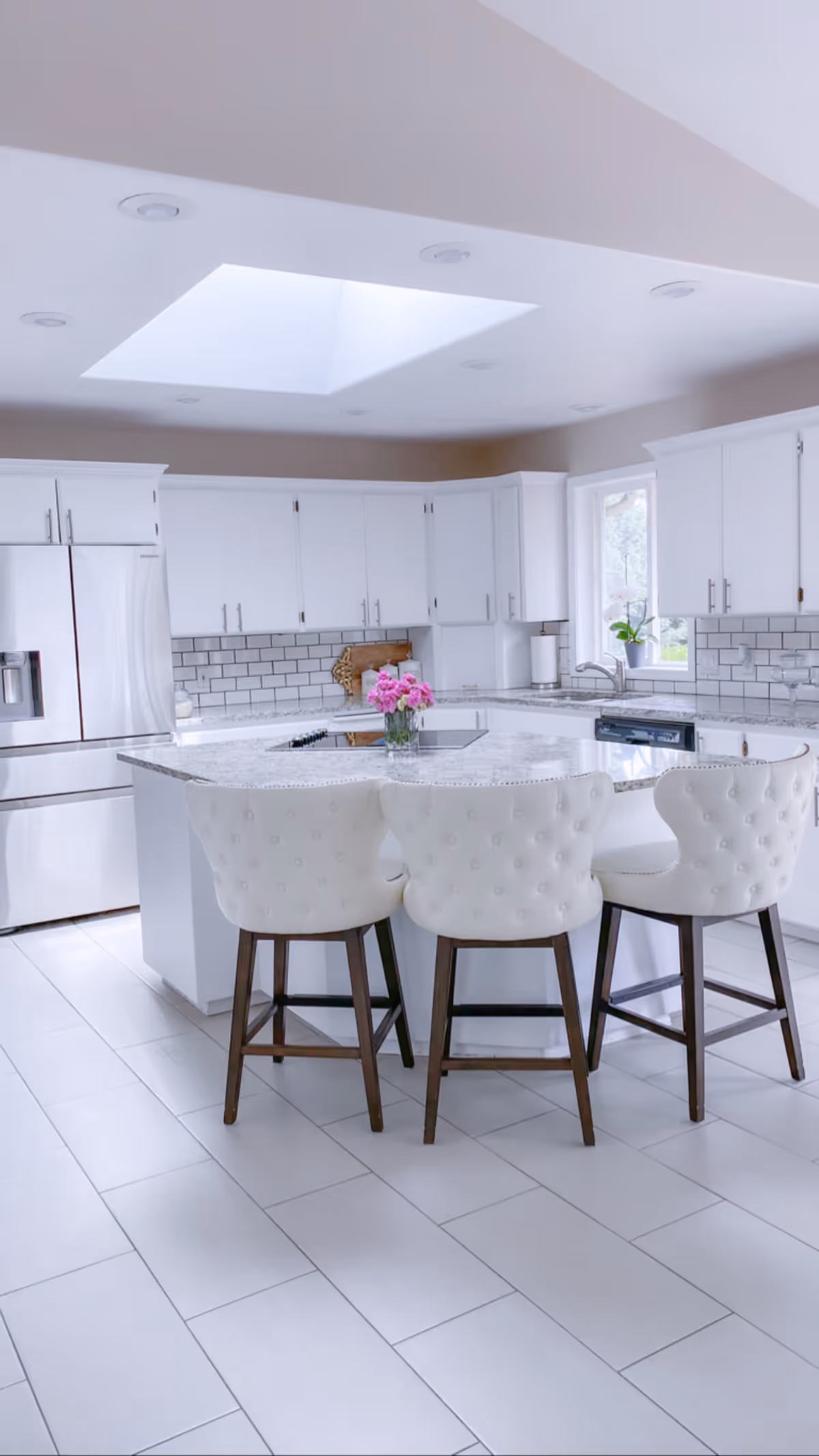 Bright modern kitchen with white cabinets, a large island with a granite countertop, three white cushioned bar stools with wooden legs, a stainless steel refrigerator, a window with a potted plant, and a skylight above providing natural light.