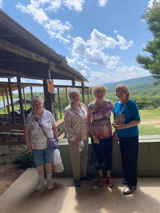 Four elderly women standing together outside near a wooden structure with a scenic view of green hills and a partly cloudy sky in the background. They are smiling and appear to be enjoying a sunny day.