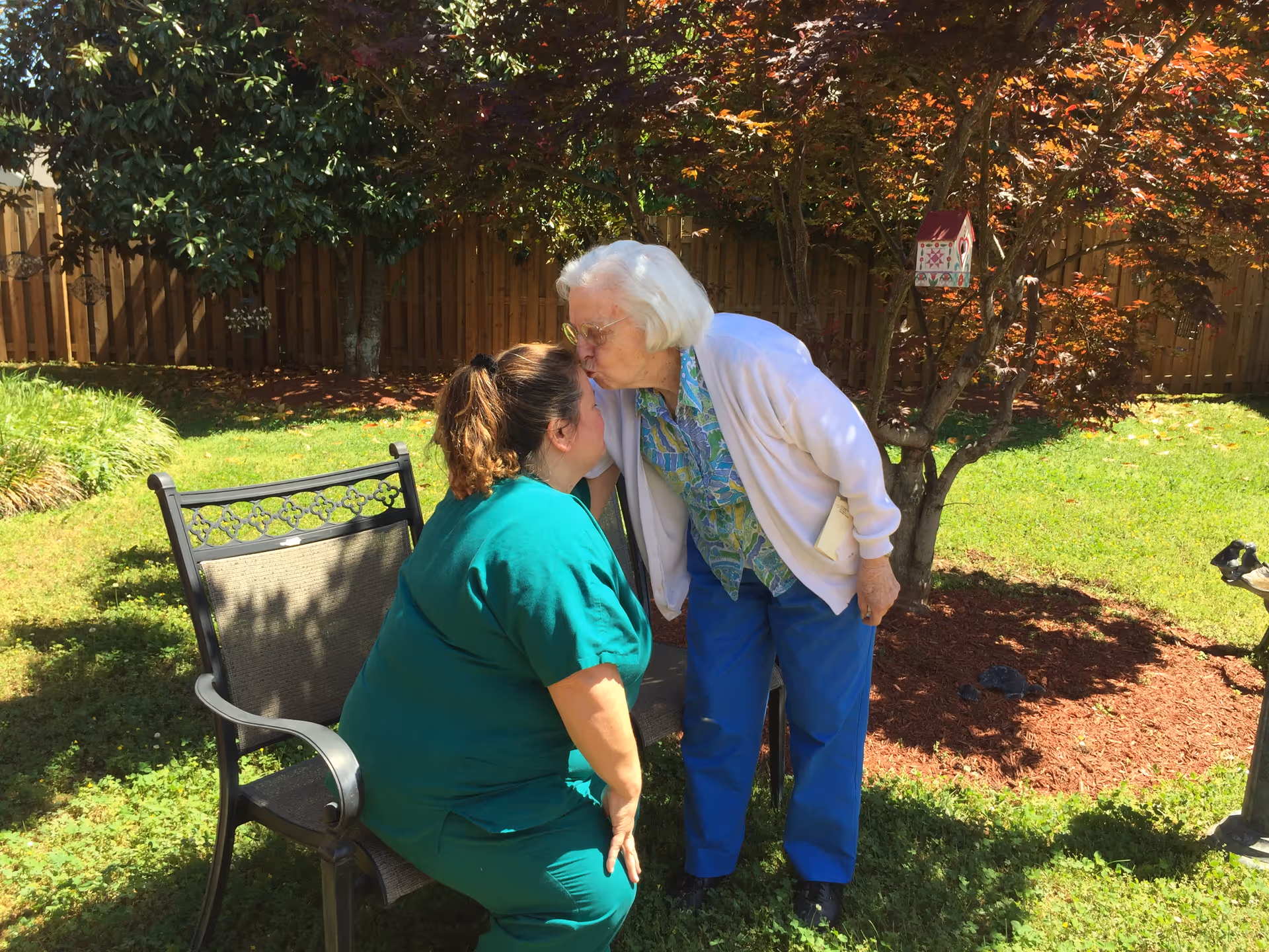 An elderly woman with white hair wearing a light purple cardigan and blue pants is leaning down to kiss a woman in green scrubs who is sitting on a metal chair in a sunny garden with green grass, trees, and a wooden fence in the background.