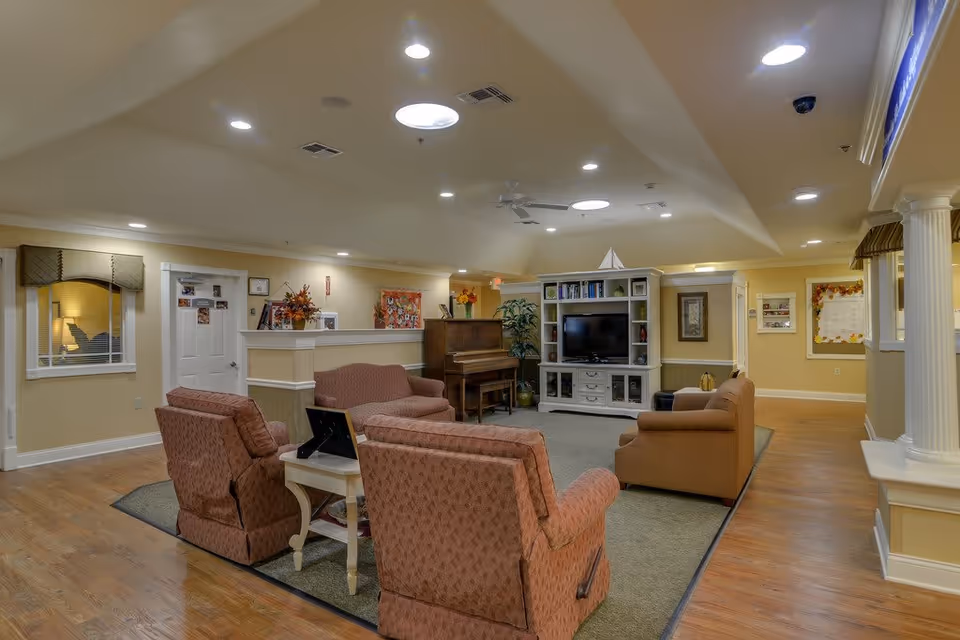 A cozy senior living facility common area with comfortable upholstered chairs and sofas arranged around a carpet. There is a white entertainment center with a TV and shelves, a piano, and decorative plants. The room has warm lighting, wood flooring, and soft yellow walls with framed artwork and decorations.