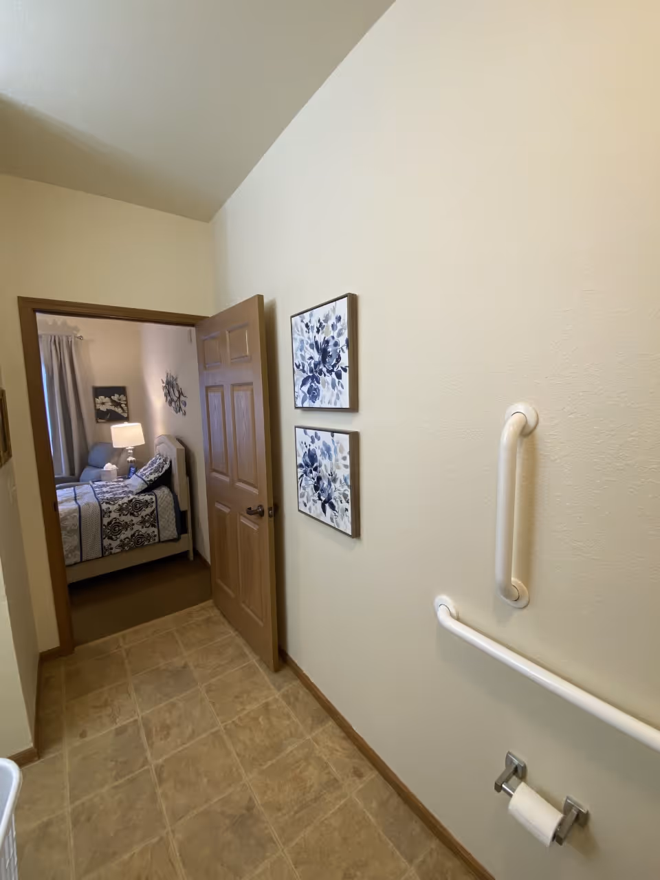 View from a bathroom looking into a bedroom. The bathroom has beige tiled floor, white walls with two grab bars and a toilet paper holder. The bedroom has a bed with patterned bedding, a nightstand with a lamp, and wall decorations including floral artwork.