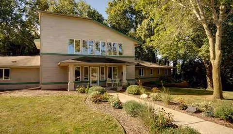 Two-story beige senior living facility with front entrance, landscaped walkway, and tall trees.