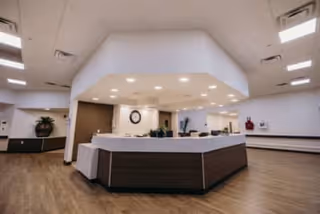 A spacious reception area with a large, modern, white and wood-paneled front desk under a white ceiling with recessed lighting. The floor is wood, and there is a plant on a bench along the wall to the left. The walls are white with some brown accents, and there is a clock on the wall behind the desk.