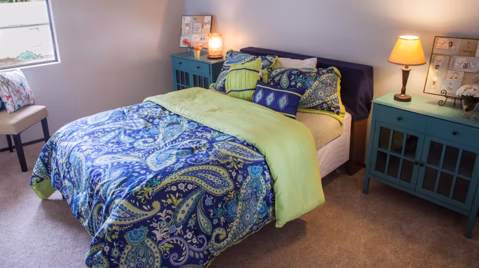 A cozy bedroom with a bed covered in a blue and green paisley patterned comforter and multiple pillows. There are two teal nightstands on either side of the bed, one with a lit lamp and decorative items. A chair with a floral cushion is placed near a window letting in natural light.