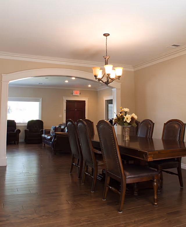 A dining room with a long wooden table and eight high-backed chairs. A vase with flowers is placed on the table. In the background, there is a living room area with leather chairs and a sofa, and a dark wooden door with an exit sign above it. The room has wooden flooring and beige walls with white trim.