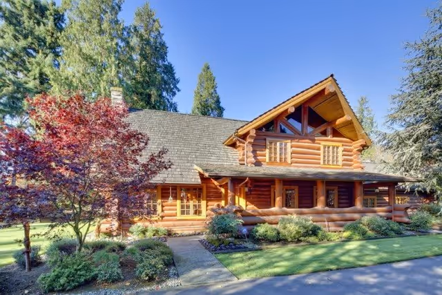 Log cabin–style building with a covered porch, landscaped front yard and walkway surrounded by trees under a clear blue sky.