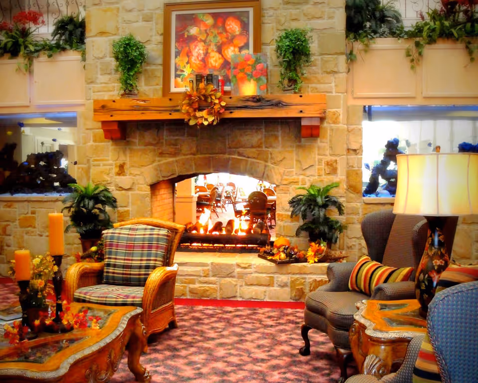 Cozy living room area with a stone fireplace in the center, decorated with a floral painting and a wreath on the wooden mantel. There are two large fish tanks built into the stone wall on either side of the fireplace. Comfortable armchairs with patterned cushions and a wooden coffee table with candles and floral decorations are arranged around the fireplace. A lamp with a floral design sits on a side table next to one of the chairs. The carpet has a red and purple pattern, and there are plants placed around the room.