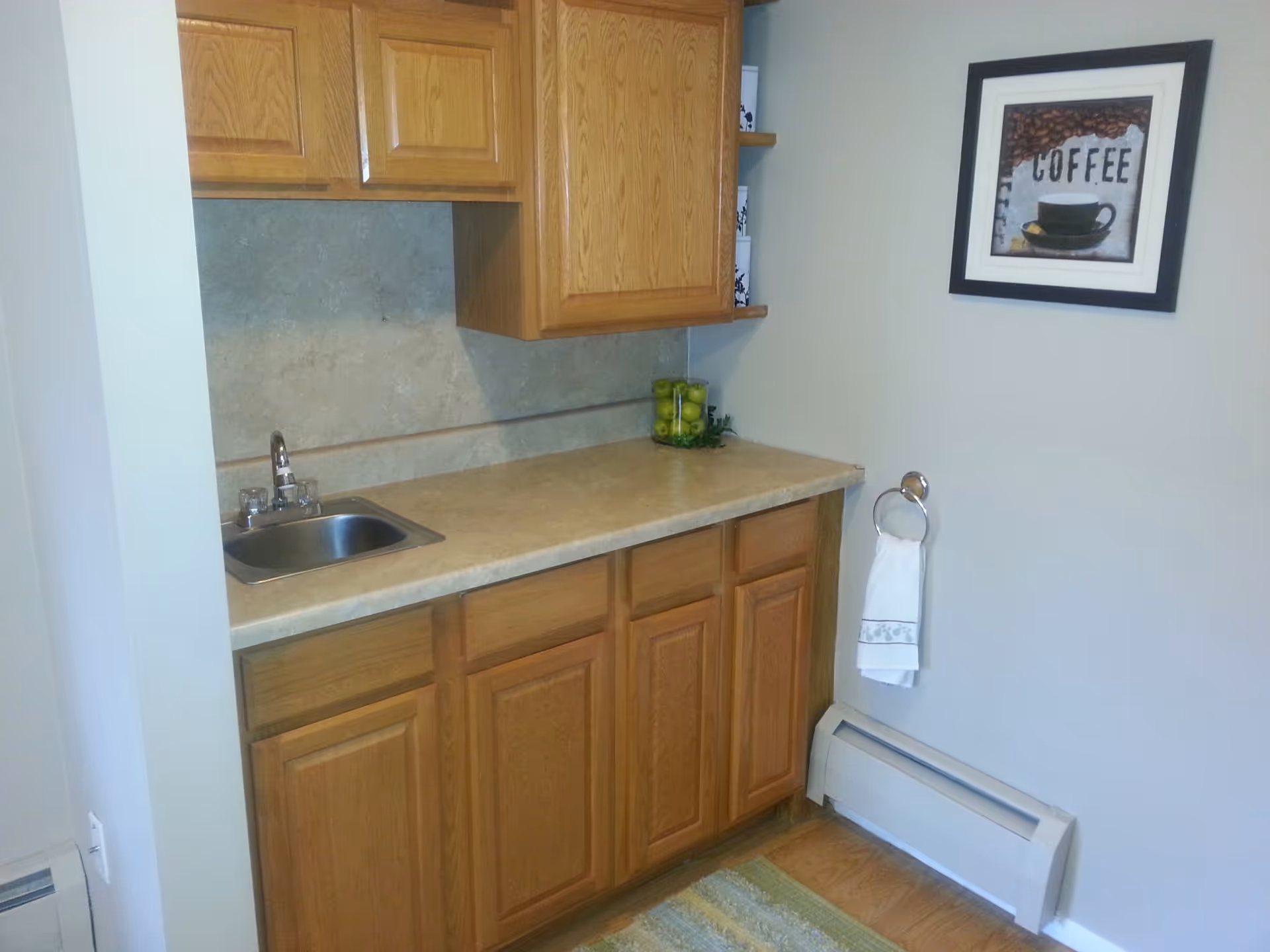 Small kitchenette area with wooden cabinets above and below a beige countertop. There is a small stainless steel sink with a faucet on the left side of the countertop. On the right side, there is a glass container filled with green apples. A white towel hangs on a ring attached to the wall, and above it is a framed picture with the word 'COFFEE' and an image of a coffee cup. The floor has a wooden finish with a green and beige rug.