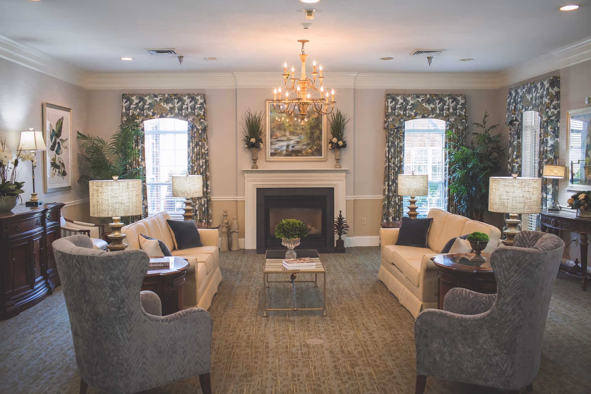 A cozy living room in Summit Hills facility featuring two beige sofas facing each other with dark throw pillows, two gray armchairs, a glass coffee table with a small green plant, and a fireplace centered on the far wall. The room is decorated with floral curtains, several table lamps, framed artwork, and indoor plants, creating a warm and inviting atmosphere.