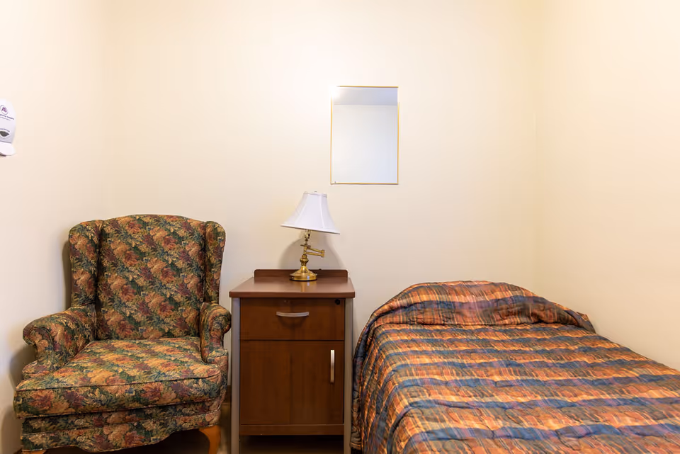 A small room with a floral upholstered armchair on the left, a wooden nightstand with a brass lamp in the center, and a single bed with a multicolored striped bedspread on the right. A small rectangular mirror is mounted on the beige wall above the nightstand.