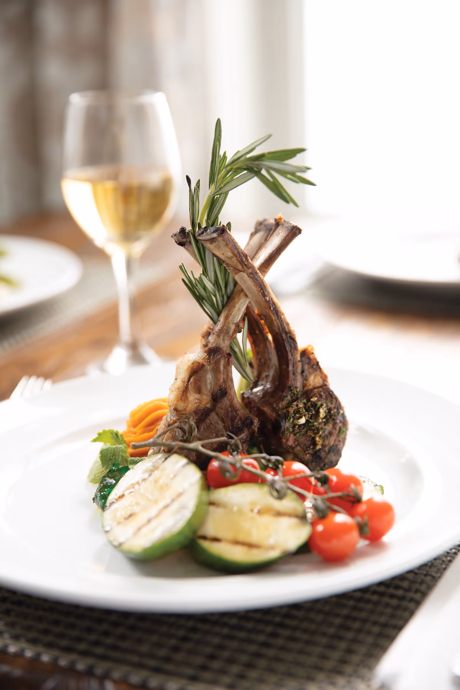 A gourmet plate featuring grilled lamb chops garnished with rosemary, accompanied by grilled zucchini slices, cherry tomatoes on the vine, and a small serving of mashed sweet potatoes. In the background, there is a glass of white wine and blurred dining table settings.